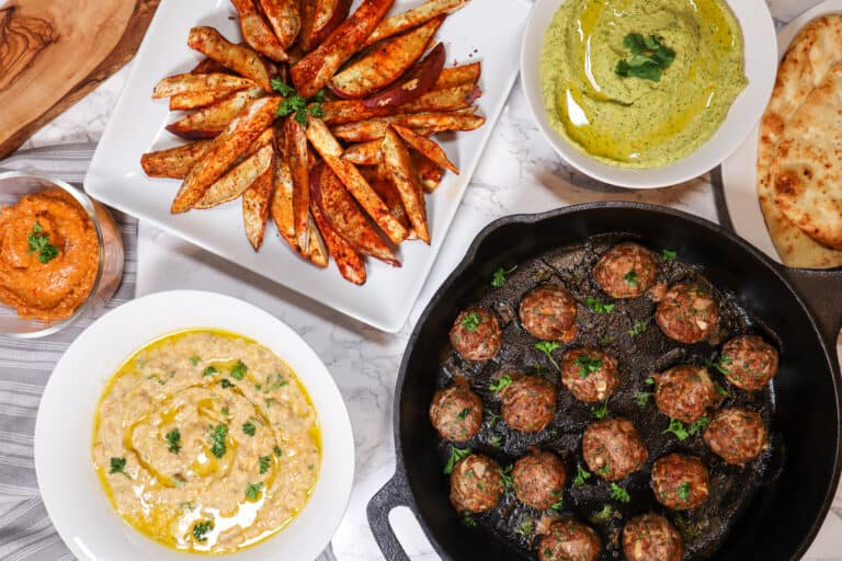 overhead shot of lamb kofta meatballs in a cast iron served with baba ganoush sweet potato fries cilantro jalapeno hummus and flat bread all are in separate bowls or plates