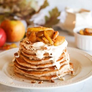 a stack of apple cinnamon pancakes on a white plate topped with maple cream cheese glaze chopped pecans and caramelized apple slices the background has apples and a ramekin of sliced apples blurred