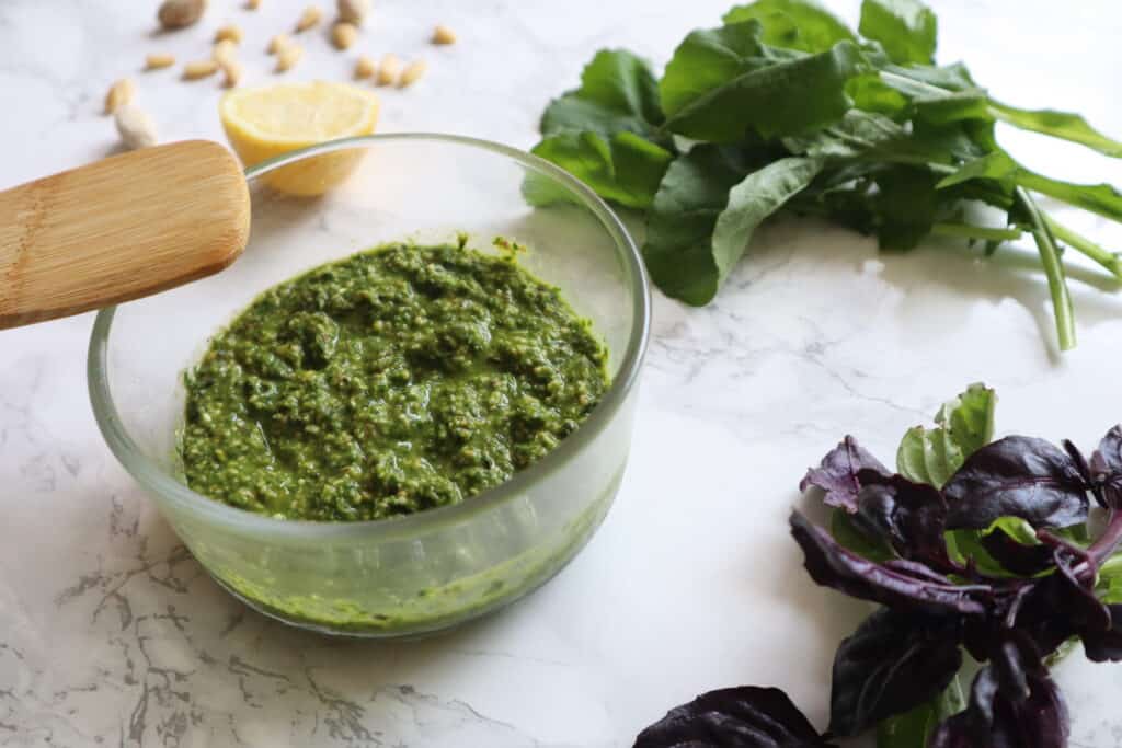 bowl of pistachio pesto with basil and lemon in the background and wooden spoon on the bowl