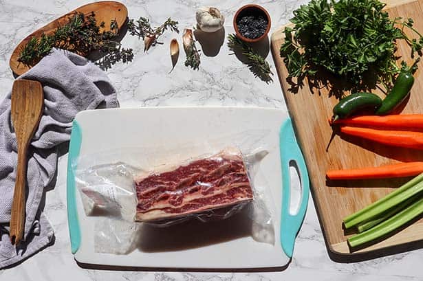 ingredients for making braised short ribs in the oven. shows are a pack of grass-fed short ribs, fresh herbs, vegetables, and garlic.