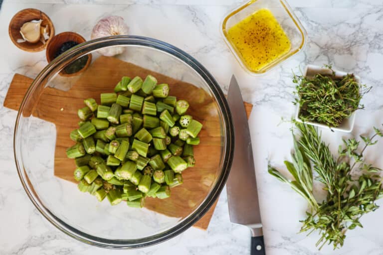 Overhead view of sliced uncooked okra on a cutting board among assorted herbs and olive oil.