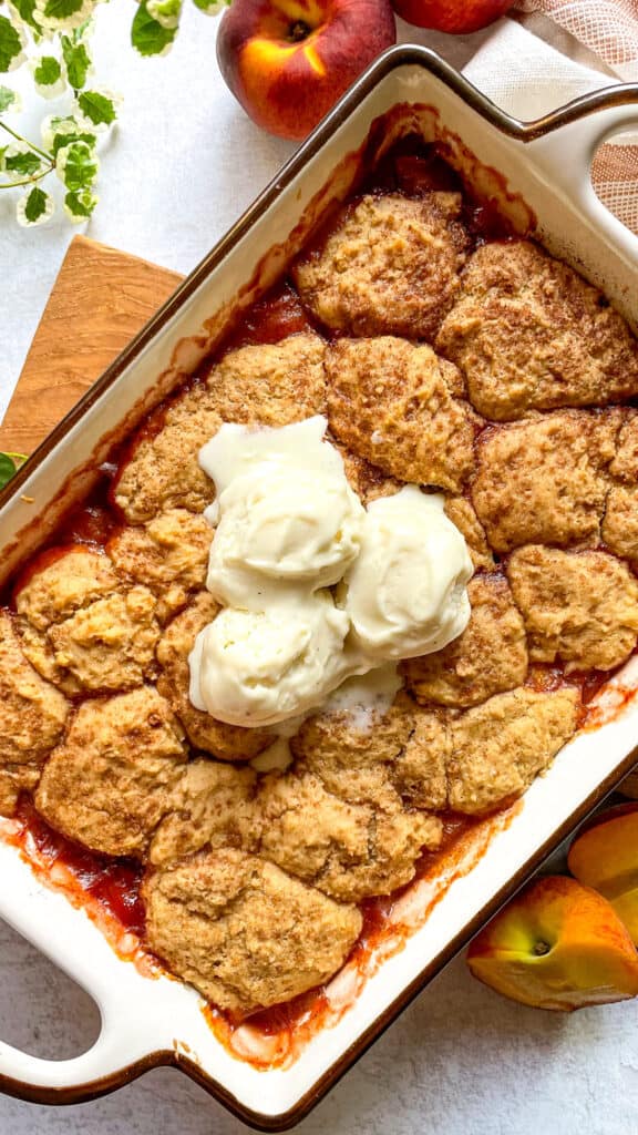 overhead shot of peach cobbler topped with vanilla ice cream in a rectangular baking dish with fresh peaches shown in the background on the site of the baking dish