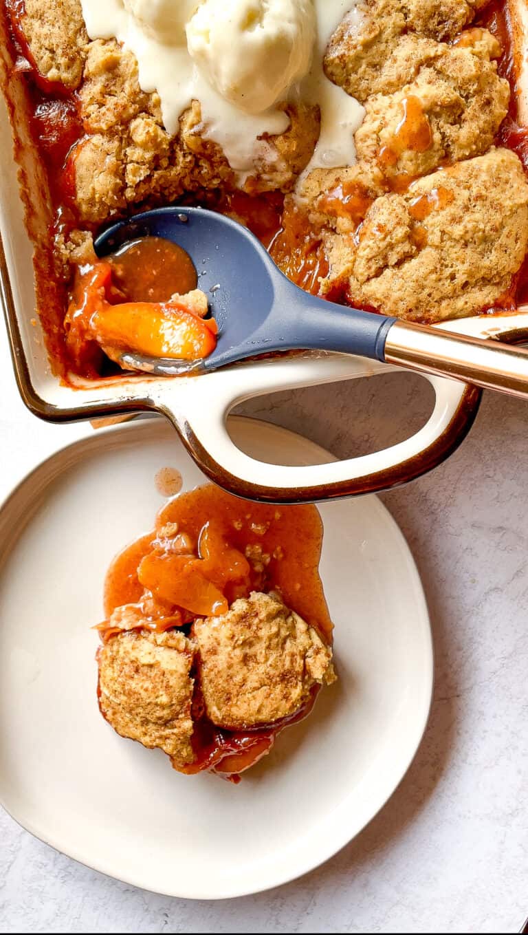 close up of peach cobbler on a plate with a baking dish of gluten free peach cobbler topped with vanilla ice cream and serving spoon in the dish