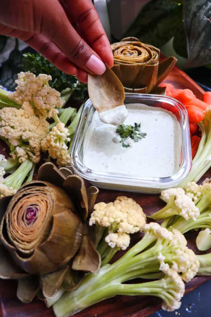 vegan ranch dressing with a vegetable platter. a hand being shown dipping artichoke into the ranch dressing