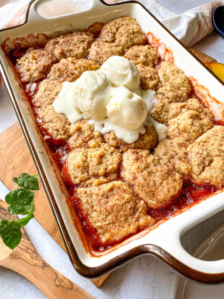 peach cobbler topped with vanilla ice cream in a rectangular baking dish with fresh peaches shown in the background on the site of the baking dish