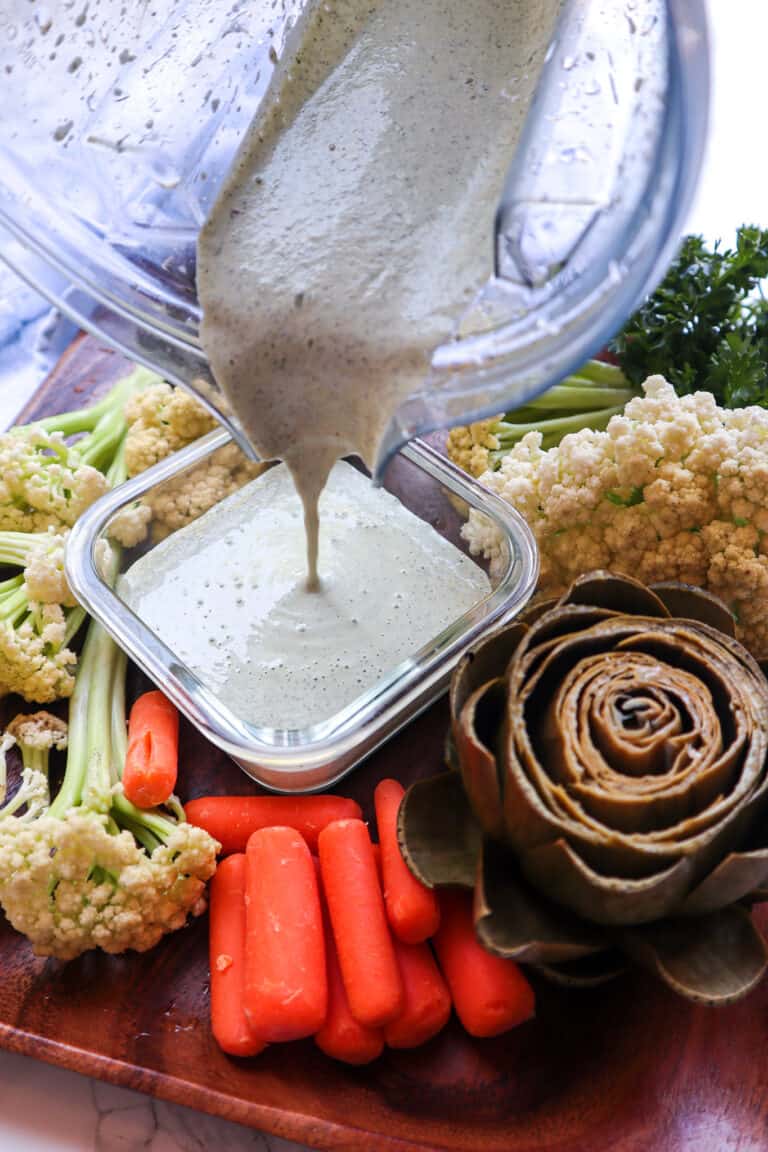 homemade vegan ranch dressing made in a blender. being poured into a small dish surrounded by vegetables