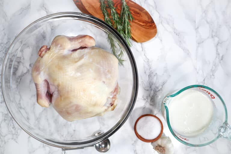 buttermilk brine for chicken and a whole chicken in a large bowl with rosemary salt and garlic on the side