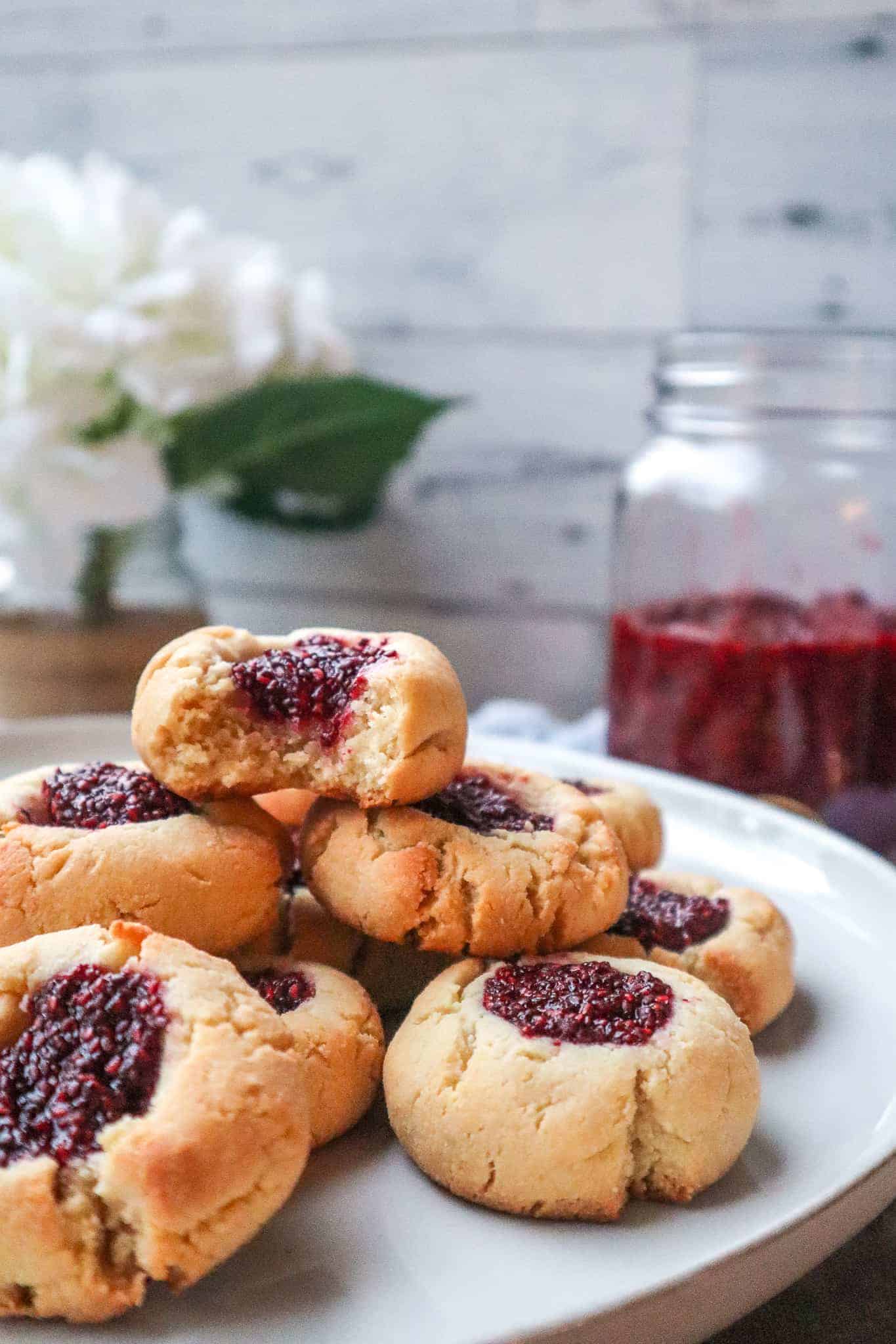 almond flour thumbprint cookies with raspberry jam on a plate stacked on top of each other with a mason jar of raspberry jam and a white flowers in the background