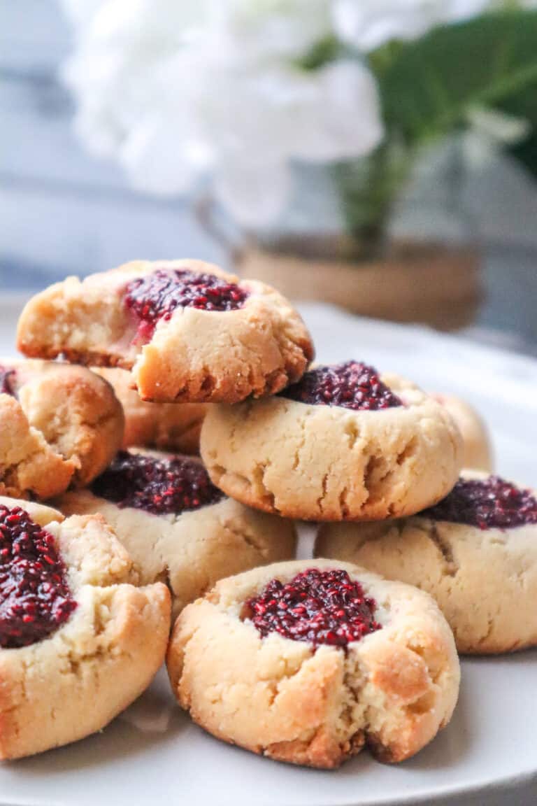 almond flour thumbprint cookies with raspberry jam on a plate stacked on top of each other with white flowers blurred in the background