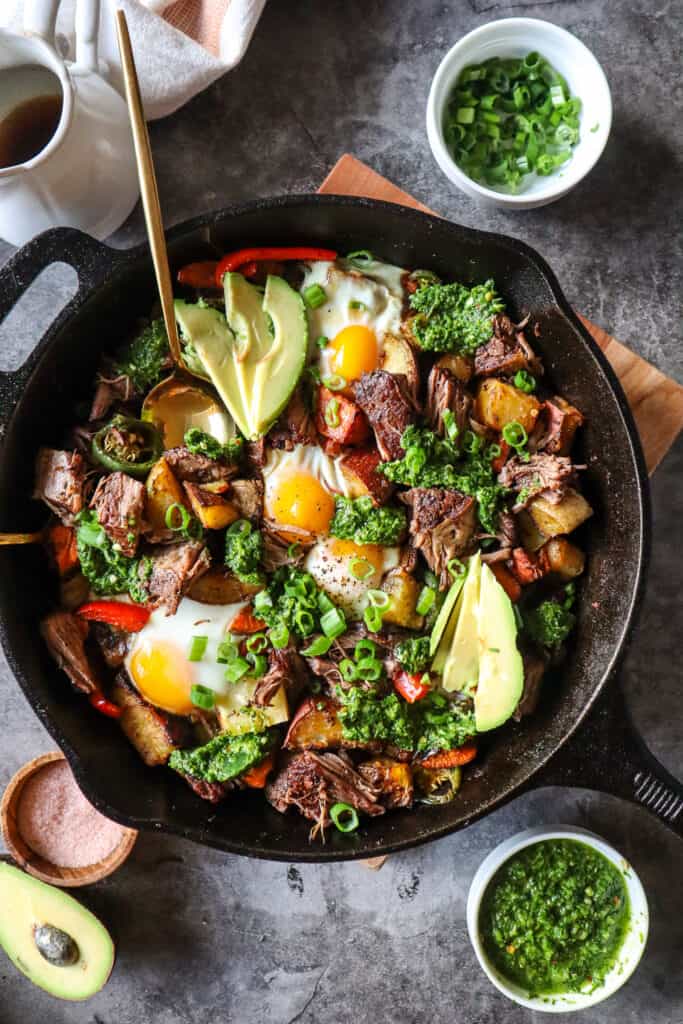 Image of beef short rib hash with sweet potatoes in a large cast iron skillet with baked eggs, topped with chimichurri, green onions, and avocado. ramekin with chimichurri in the top left corner, chopped green onions in the bottom right corner, a small bowl, and half of avocado in the bottom left corner. 