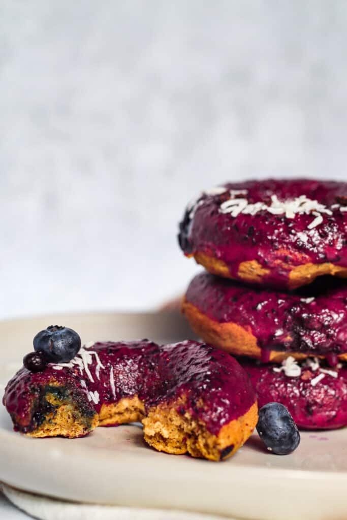 close up image of bitten blueberry donut to show texture with stack of vegan blueberry baked donuts shown