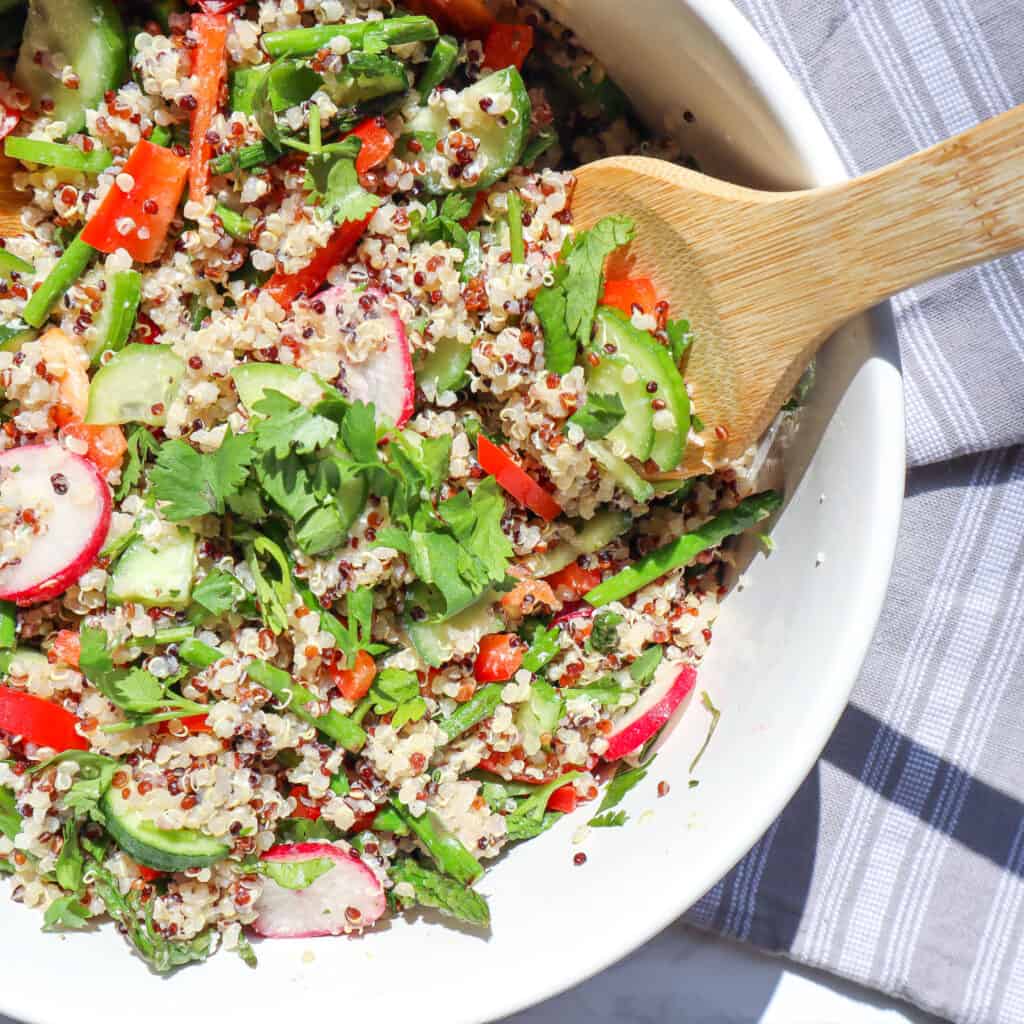 a mix of cooked quinoa asparagus cucumber red pepper radishes cilantro and goat cheese in a large bowl with one wooden spoons in the bowl