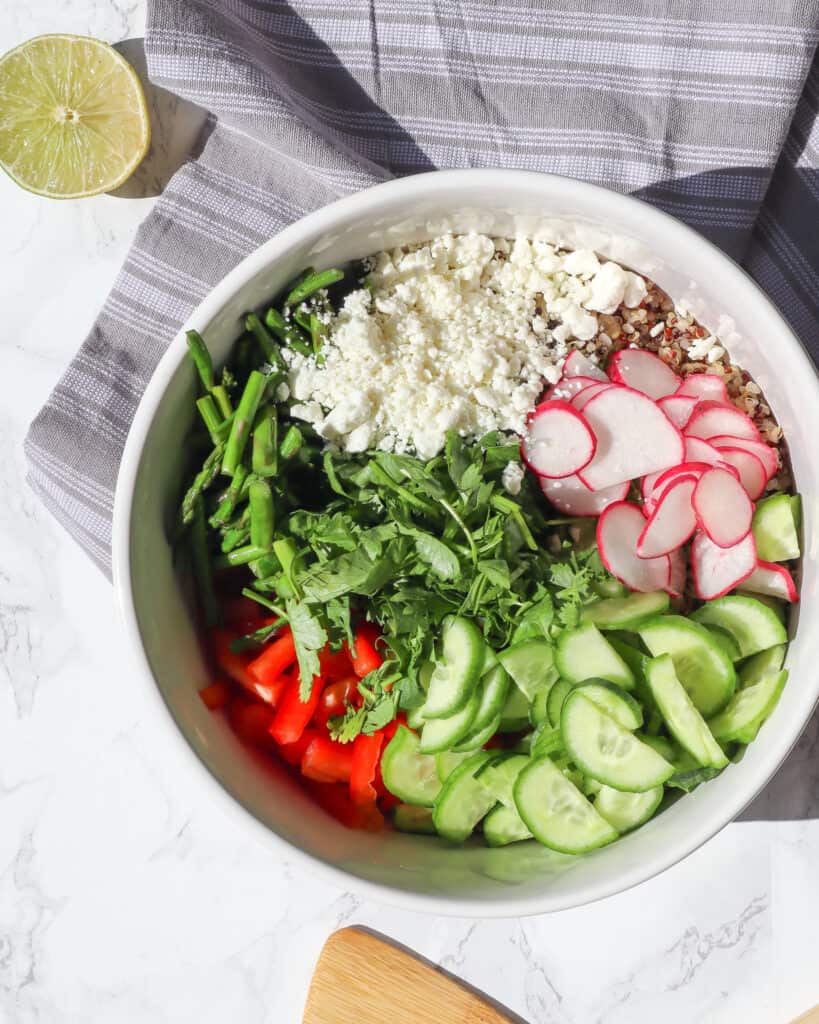 cooked quinoa asparagus cucumber red pepper radishes cilantro and goat cheese in a large bowl with a half lime in the top left corner