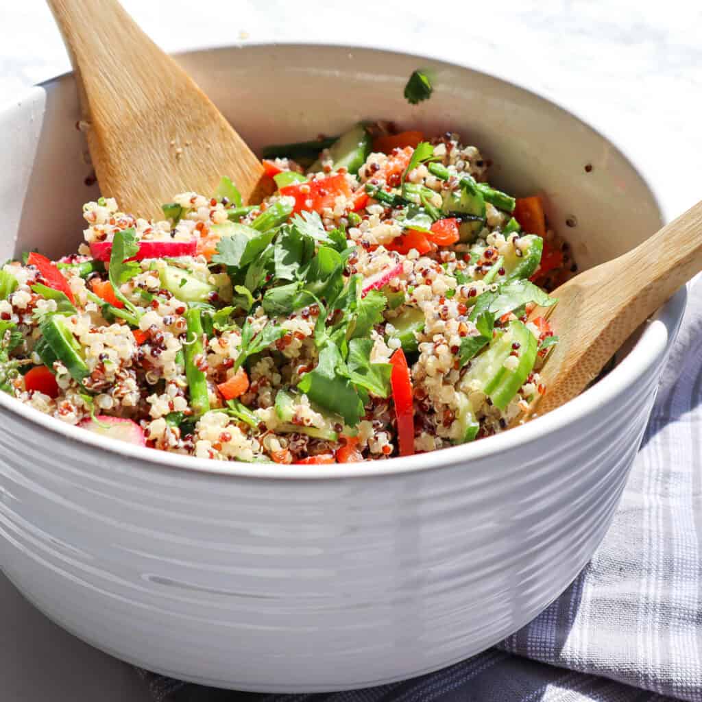 a mix of cooked quinoa asparagus cucumber red pepper radishes cilantro and goat cheese in a large bowl with two wooden spoons in the bowl