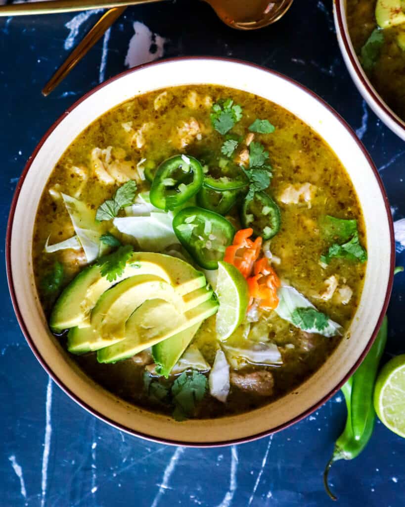 Bowl of Pork Pozole Verde with Toppings of avocado, jalapeno, lime, cabbage, and habanero