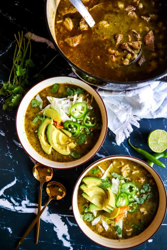 2 bowls of Pork Pozole Verde with Toppings of avocado, jalapeno, lime, cabbage, and habanero