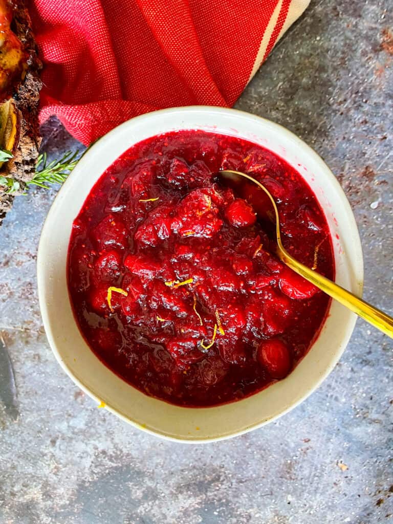 Orange Cranberry Sauce in a bowl with a gold spoon. Red cloth and persimmon in top left corner of image