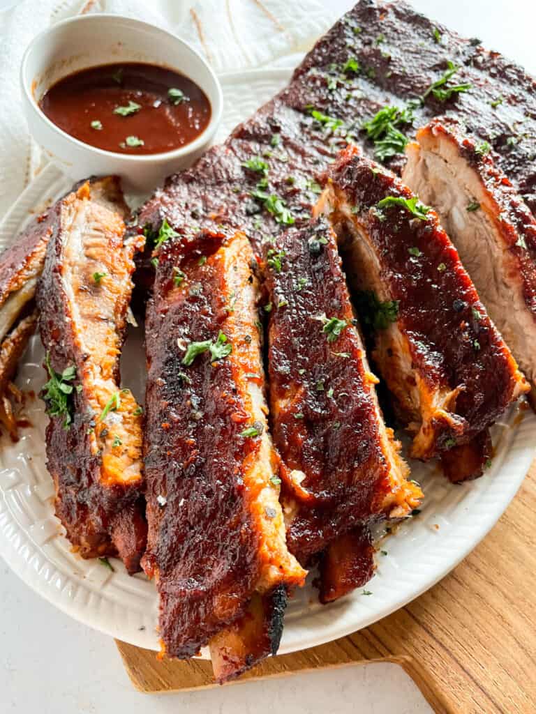 A white platter full of Instant Pot Ribs, sliced with a small bowl of homemade barbecue sauce in the upper left corner.