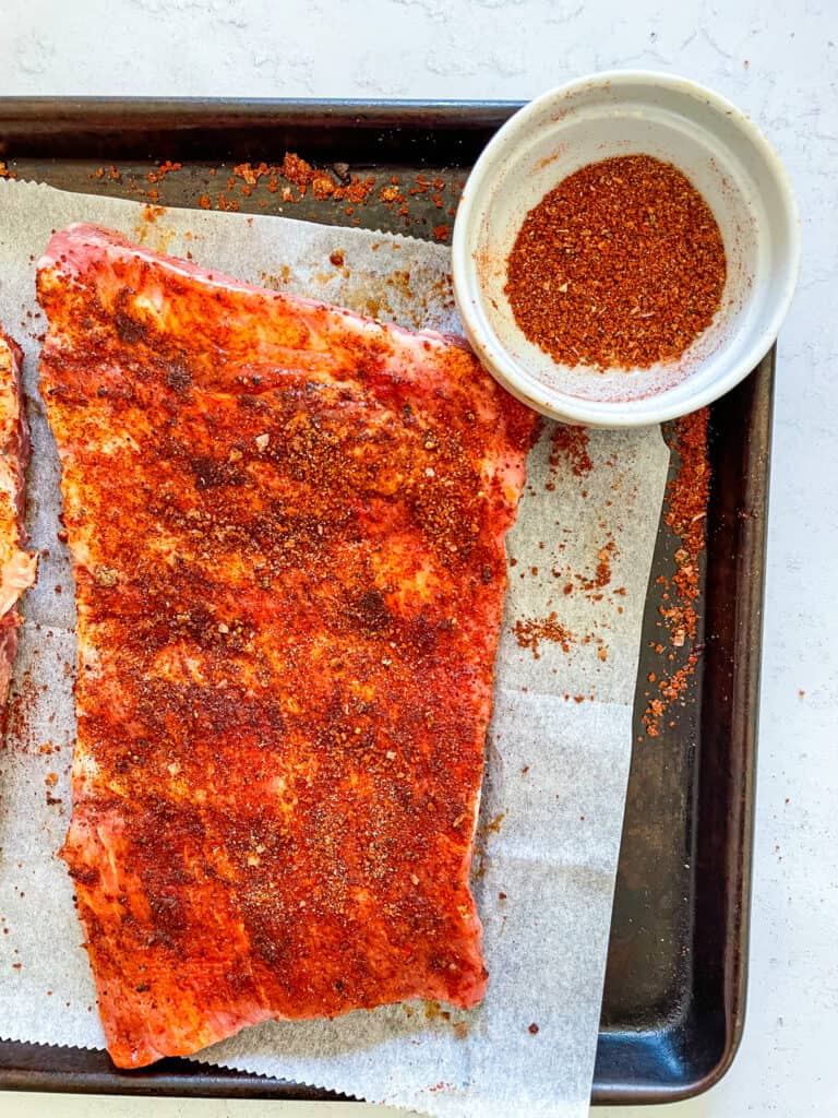 An overhead view of baby back ribs covered in dry rub with a small bowl of extra dry rub on the right side.