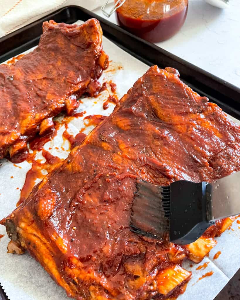 Two slabs of Instant Pot baby back ribs on parchment paper on a baking tray being coated in homemade barbecue sauce.
