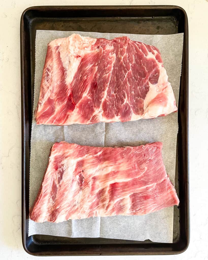 Overhead view of two slabs of raw baby back ribs on a sheet of parchment paper on a baking sheet.