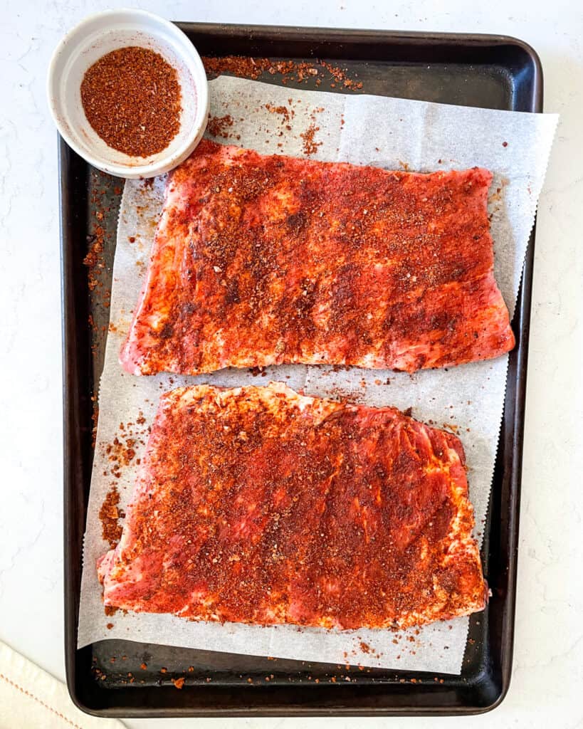 Overhead view of two slabs of raw baby back ribs coated in dry rub with a small bowl of dry rub in the upper left corner. The ribs are on a sheet of parchment paper on a baking sheet.
