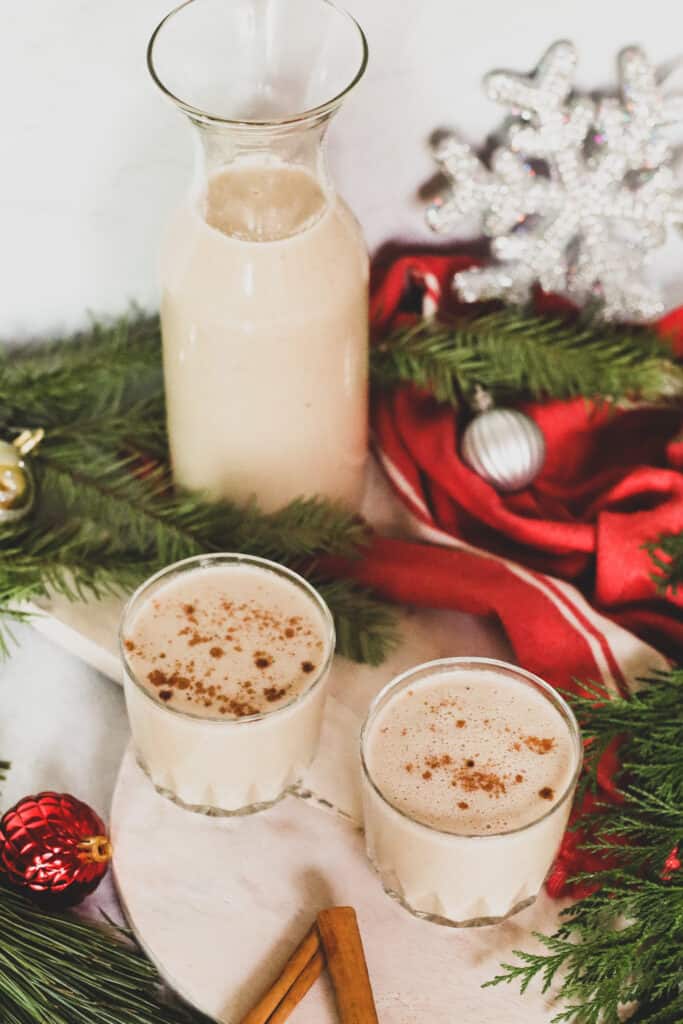 two gluten free dairy free eggnog drinks with cinnamon stick next to the drink and decorative ornaments and christmas garland