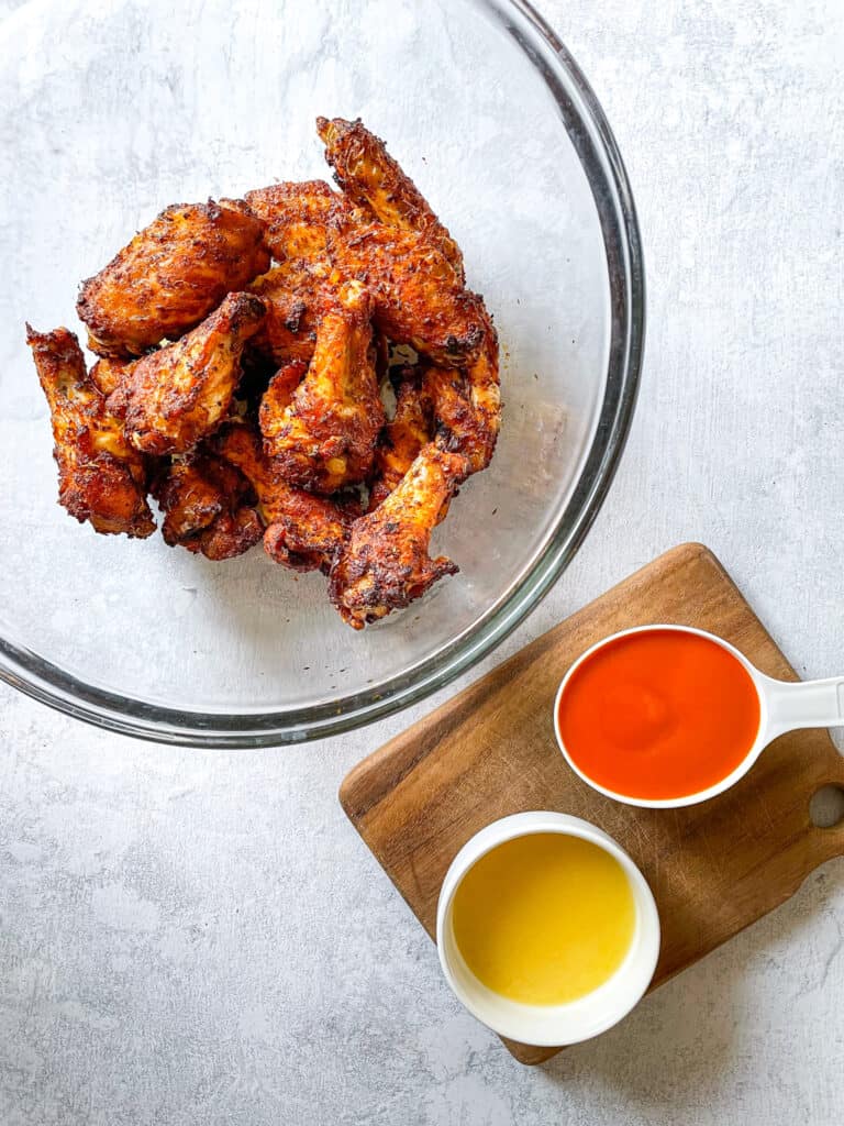 air fryer wings in a bowl next to buffalo sauce in a measuring cup and butter in a ramekin