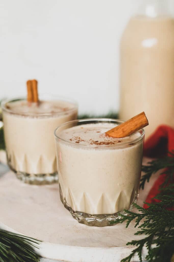 two gluten free dairy free eggnog drinks with cinnamon stick next to the drink and decorative ornaments