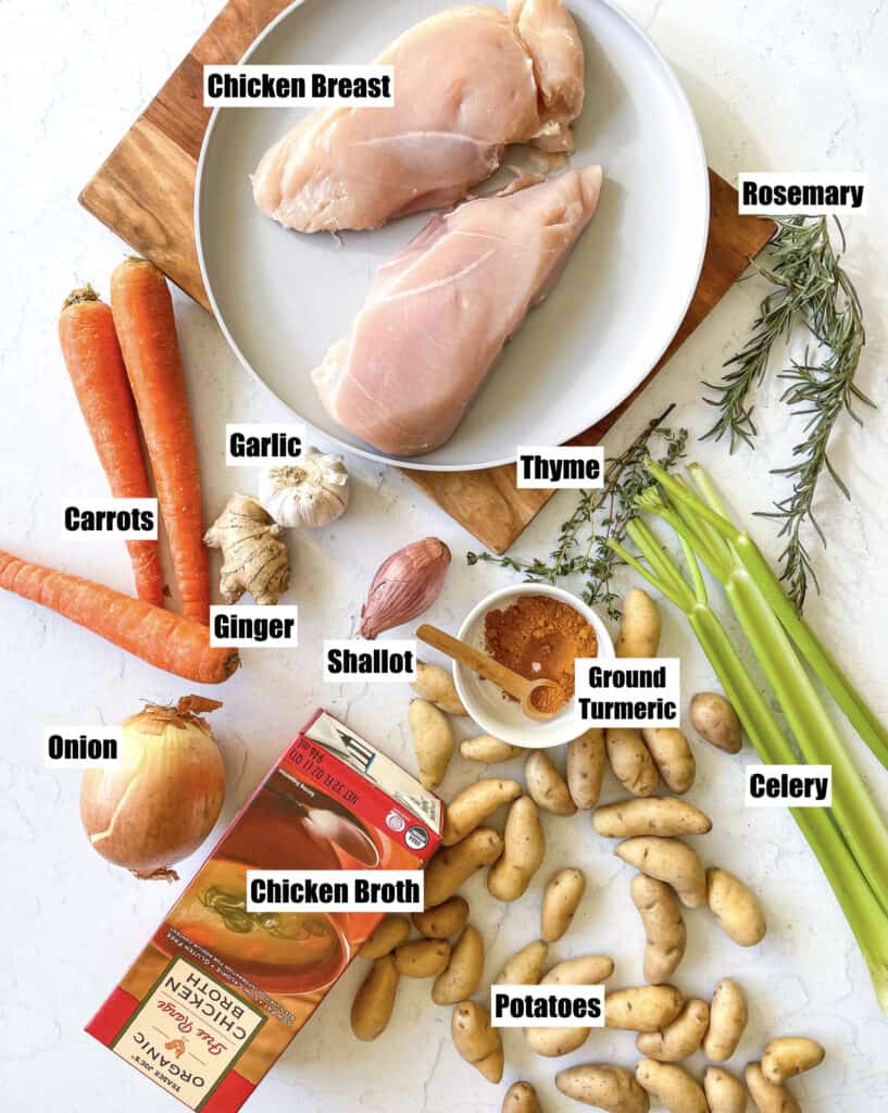 ingredients needed. shown are 2 raw chicken breast on a plate, 3 carrots, a whole onion, a whole shallot, a whole garlic bulb, ginger, ground turmeric, rosemary, thyme, potatoes, 3 celery stalks, and a box of chicken broth