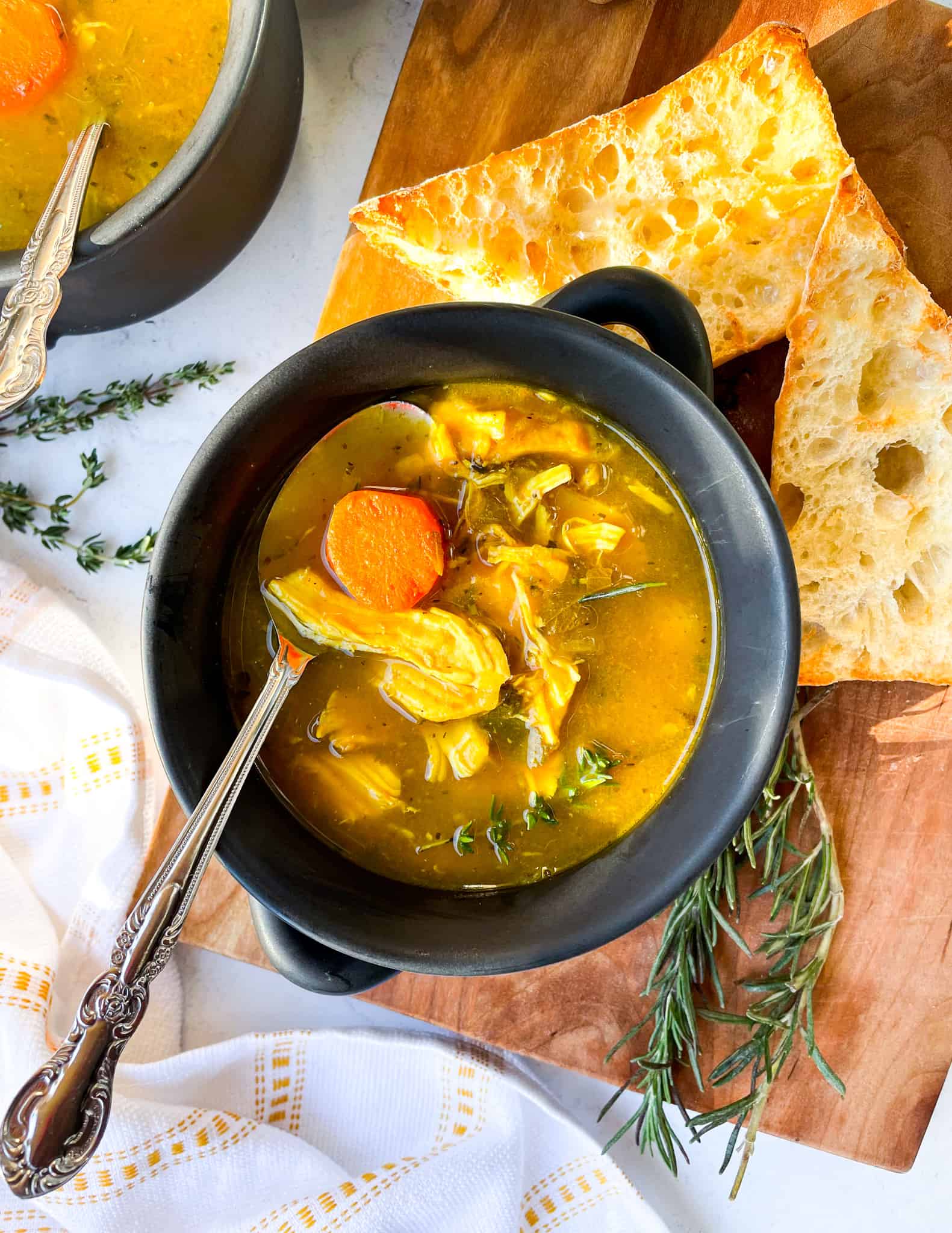 bowl of of chicken soup with a spoon in bowl showing shredded chicken and broth. slice of bread next to bowl along with fresh herbs and a cloth