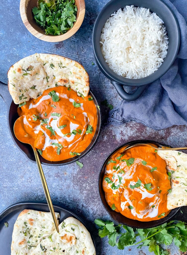 dairy free butter chicken plated in two small cast irons. shown with a side of basmati rice and naan. decorative cloth in background with cilantro leaves next to dish