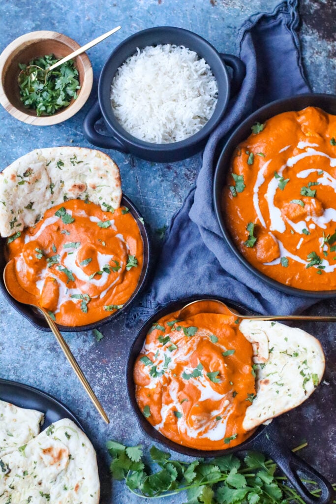 dairy free butter chicken plated in two small cast irons. shown with a side of basmati rice and naan. decorative cloth in background with cilantro leaves next to dish