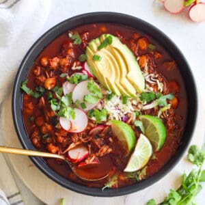 Chicken Pozole Rojo in a large bowl with toppings of lime, radish, avocado, cilantro, and green onion
