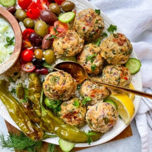 greek chicken meatballs plated with peppers, olives, rice, sliced cucumbers, lemon, and a small bowl of homemade tzatziki sauce