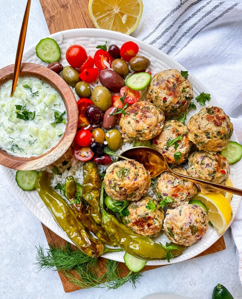 greek chicken meatballs plated with peppers, olives, rice, sliced cucumbers, lemon, and a small bowl of homemade tzatziki sauce