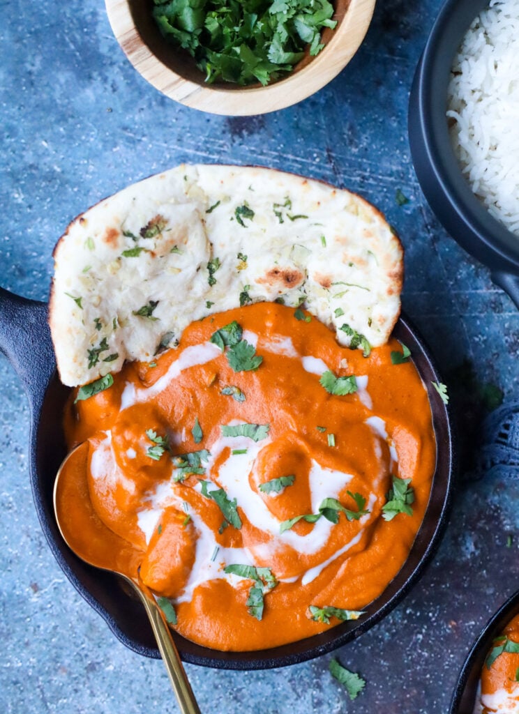 close up of dairy free butter chicken garnished with coconut cream and fresh cilantro leaves and a side of naan. rice in top right corner in background