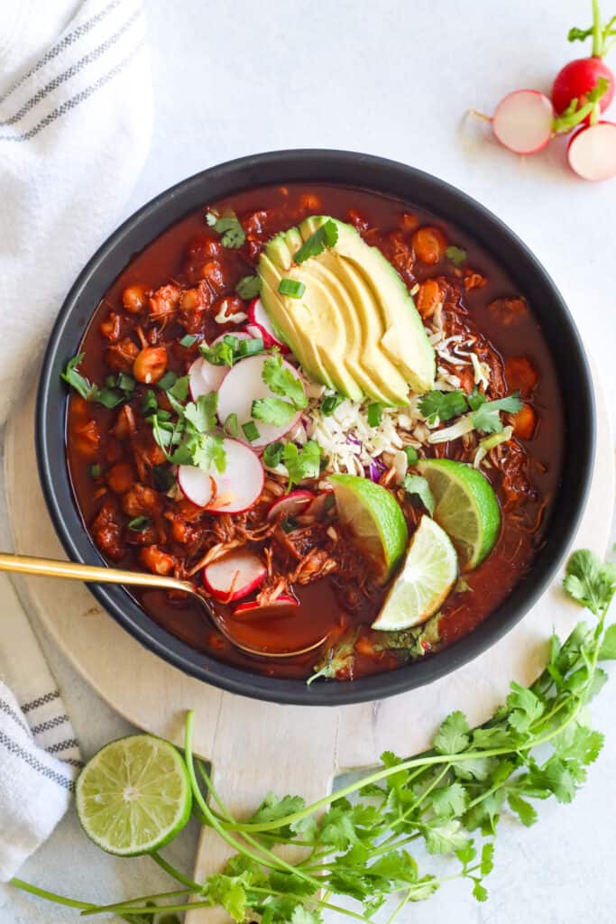 Chicken Pozole Rojo in a large bowl with toppings of lime, radish, avocado, cilantro, and green onion