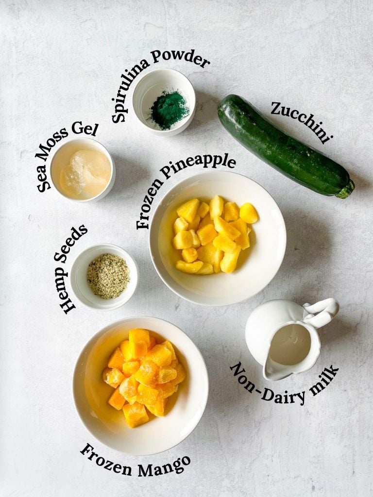 ingredients needed to make a spirulina smoothie: frozen mango, frozen pineapple, non-dairy milk, hemp seeds, raw zucchini, spirulina powder, sea moss gel