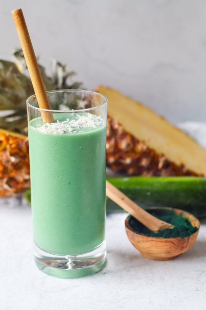 1 spirulina smoothie with spirulina powder, sliced pineapple, and zucchini in the background