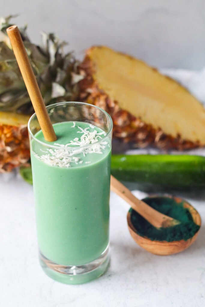 1 spirulina smoothie with spirulina powder, sliced pineapple, and zucchini in the background