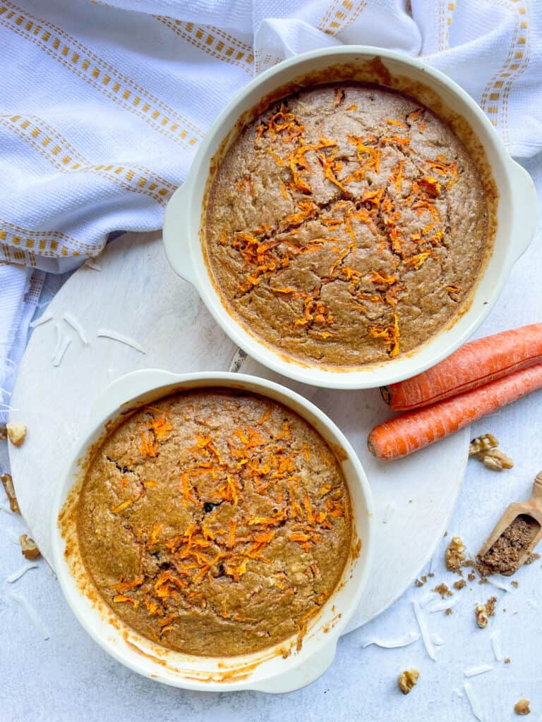 two ramekins full of baked oatmeal with carrots with various ingredients scattered around including carrot sticks, walnuts, brown sugar and shredded coconut