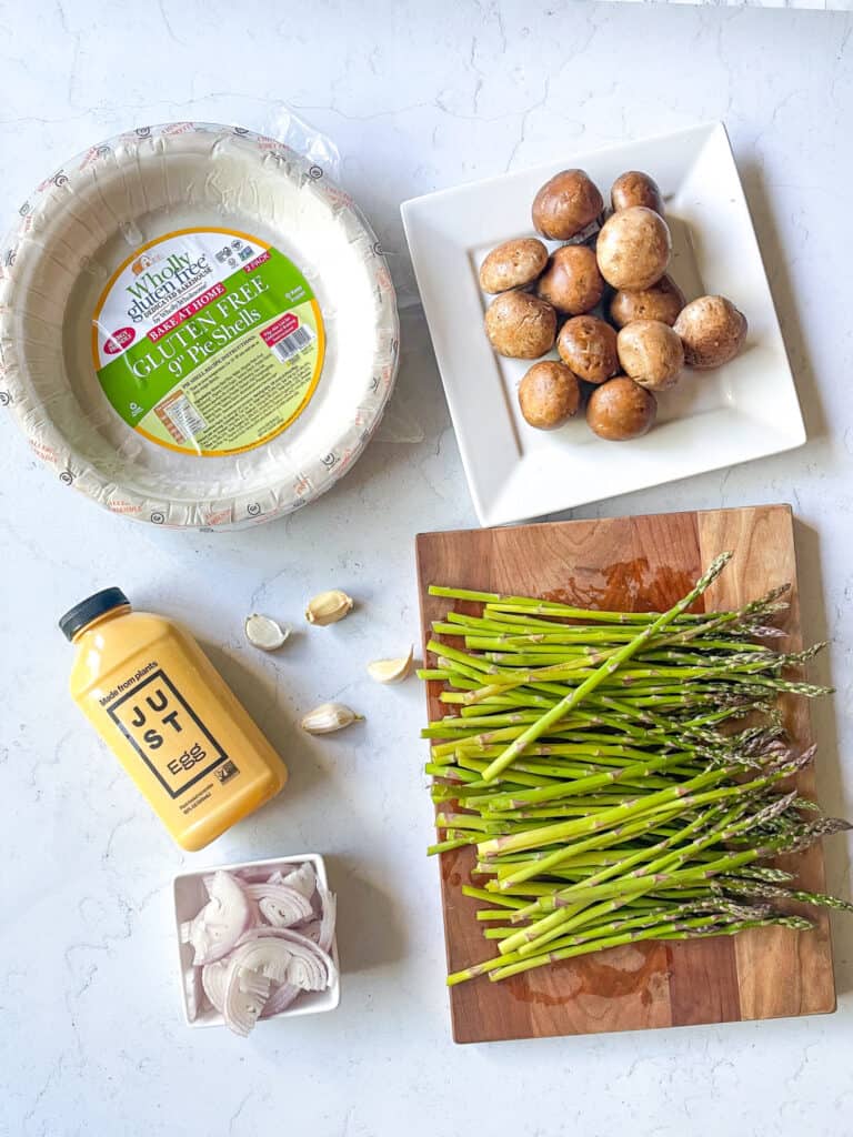 Ingredients needed to make just egg quiche shown are a gluten-free crust crimini mushrooms, bottle of just egg, garlic, shallots, and asparagus