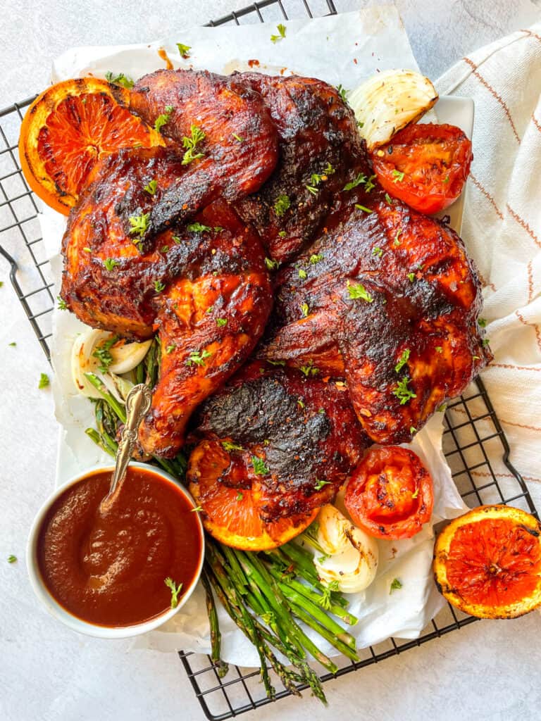 a whole spatchcocked chicken with a side of barbecue sauced, sliced oranges, grilled tomatoes, onions and asparagus 