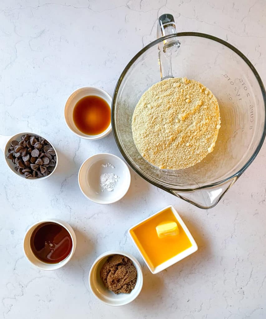 An overhead view of the ingredients needed to make eggless chocolate chip cookie dough including almond flour, baking powder, butter, maple syrup, brown sugar, vanilla extract, chocolate chips and salt.