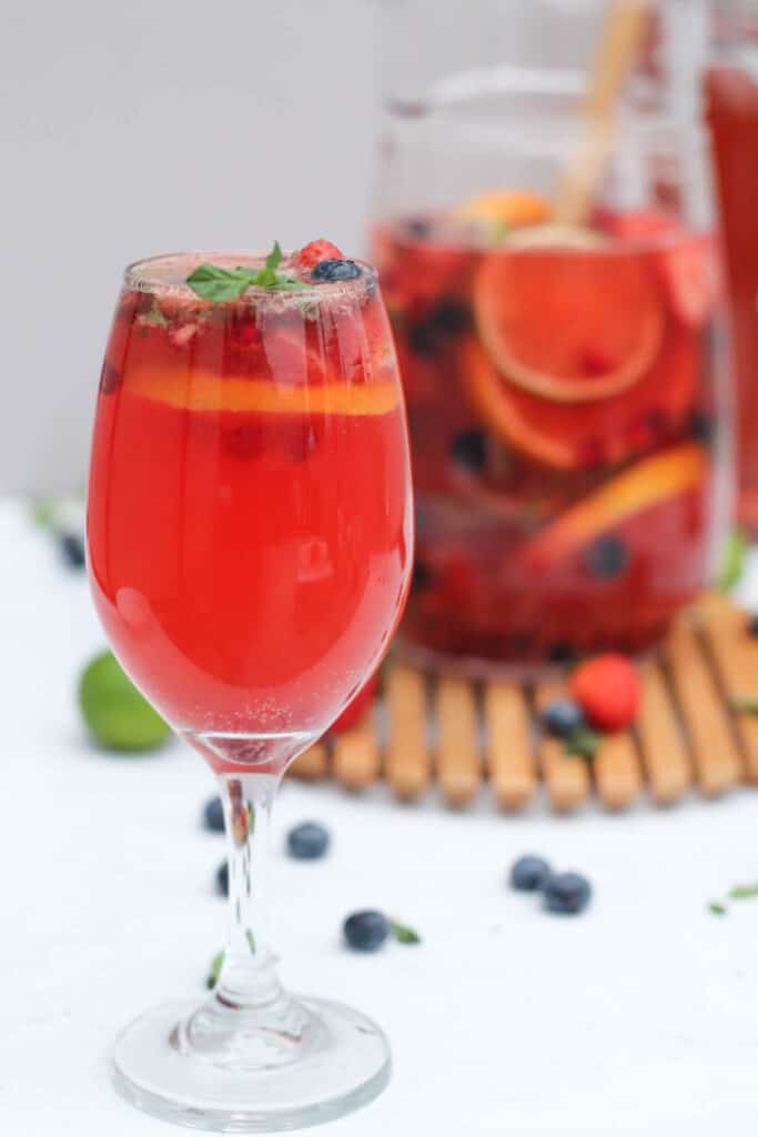 a close up of a glass of rose sangria with fresh fruit and a large pitcher of sangria made with rose in the background