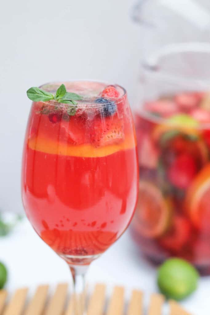 a close up of a glass of rose sangria with fresh fruit and a large pitcher of sangria made with rose in the background