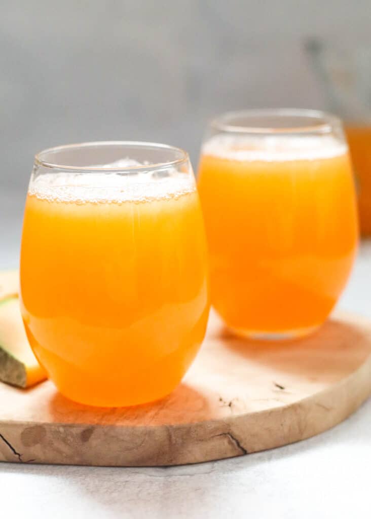 two glasses of cantaloupe juice on a wooden board with a slice of cantaloupe next to the glasses