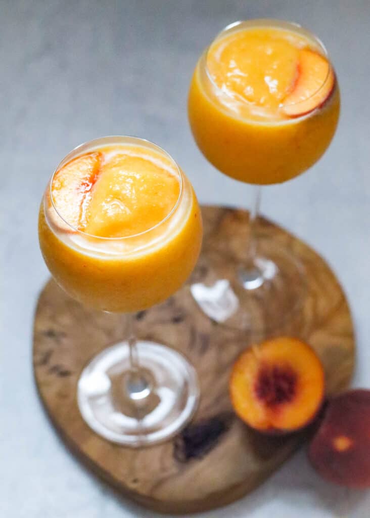 two peach wine slushies on a wooden board with fresh peaches in background