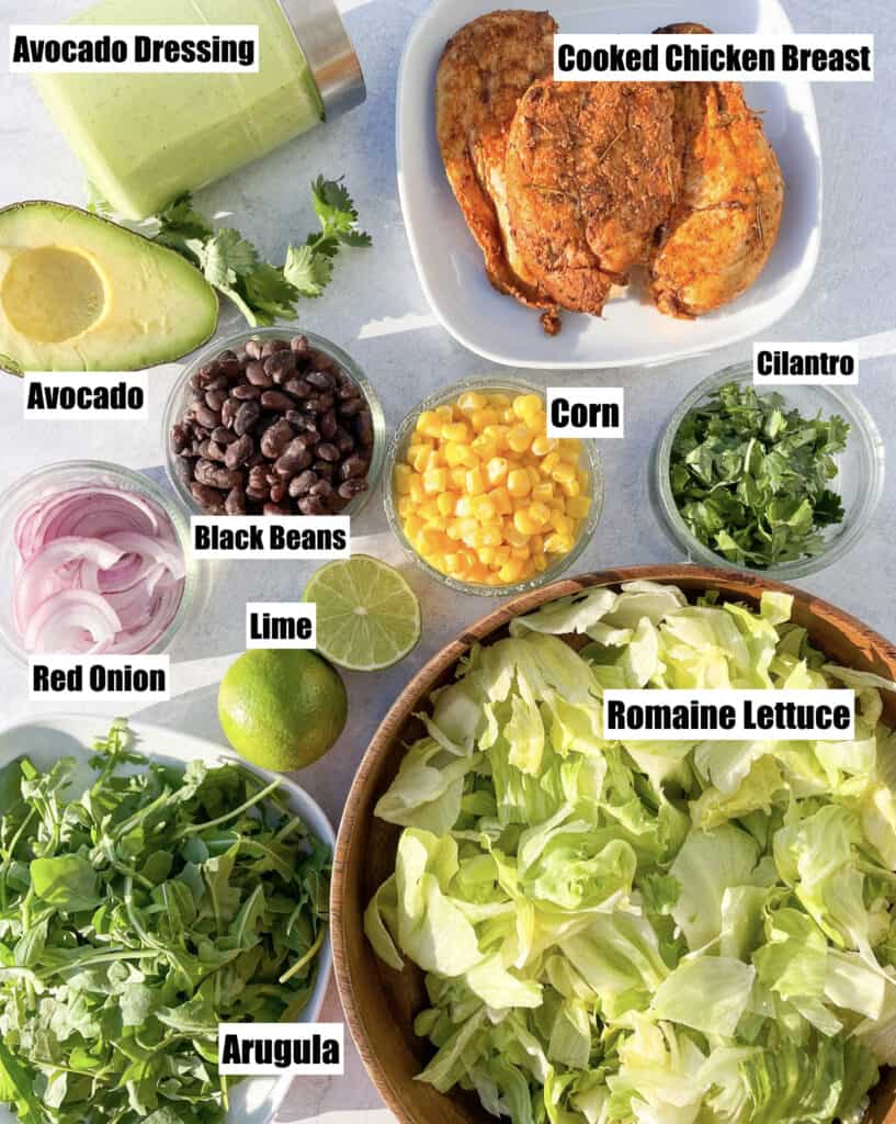 ingredients to make a southwest salad are shown there is avocado cooked chicken cilantro corn black beans lime romaine lettuce arugula red onion and avocado dressing shown 
